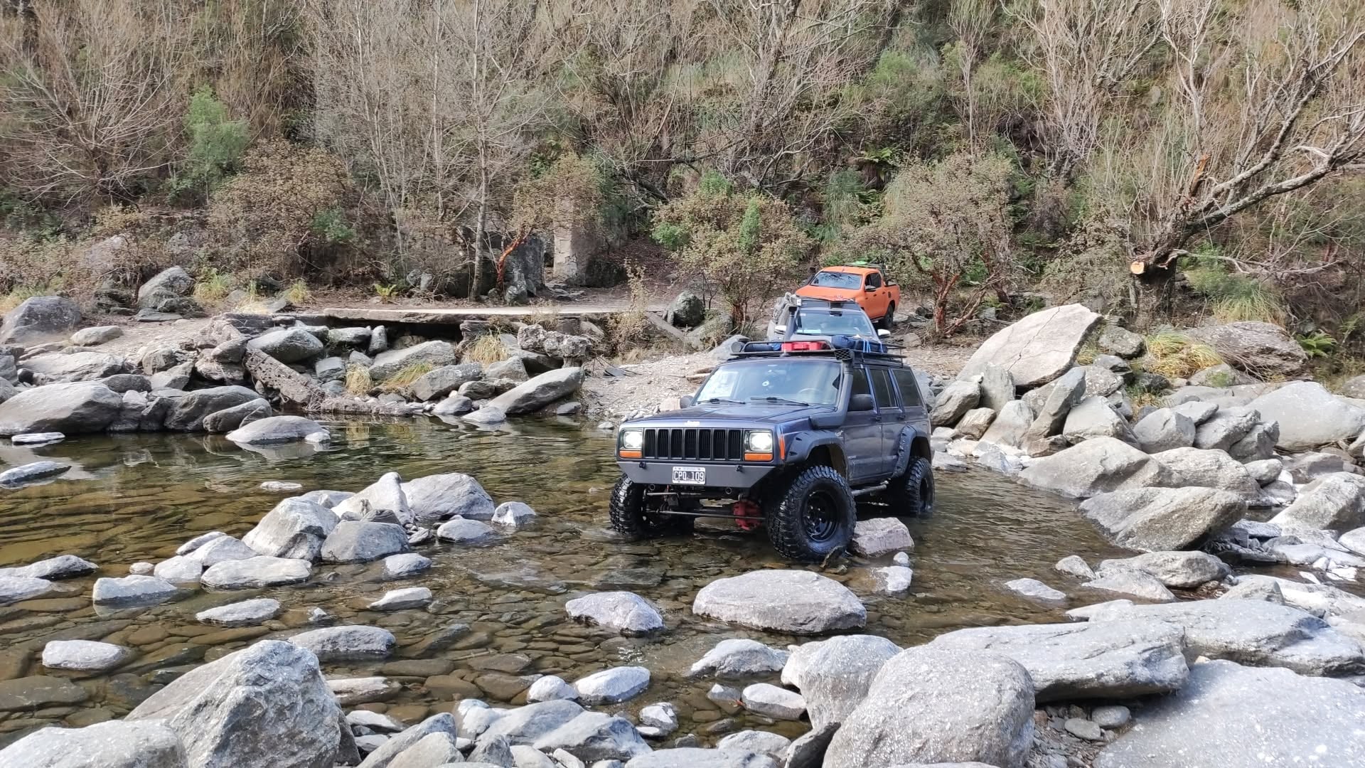 Jeep Cherokee y caravana de camionetas cruzando un arroyo entre rocas